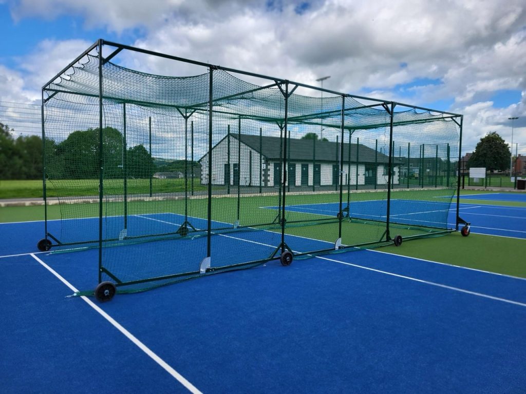 Angled front view of a cricket cage with green frame and netting, set up across a blue and green tennis surface.