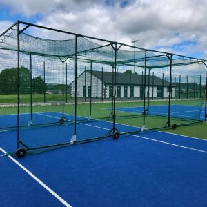 Angled front view of a cricket cage with green frame and netting, set up across a blue and green tennis surface.