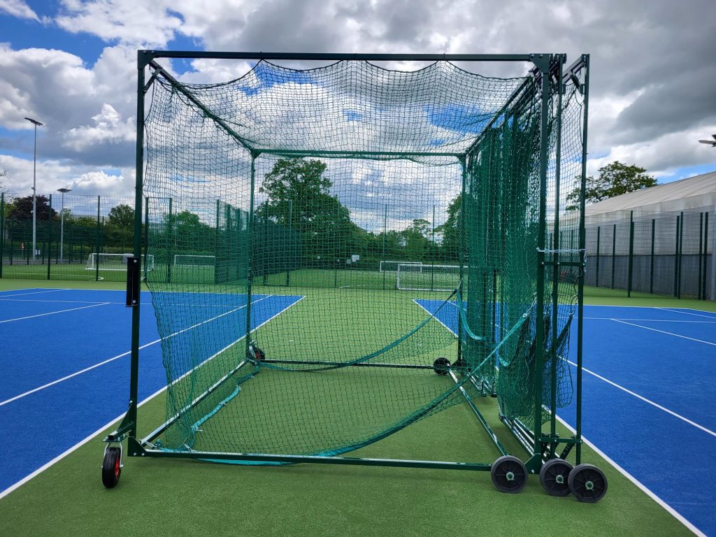 Folded green cricket batting cage with wheels positioned on a blue tennis court, showing Batting Cages Australia’s portable and compact design.